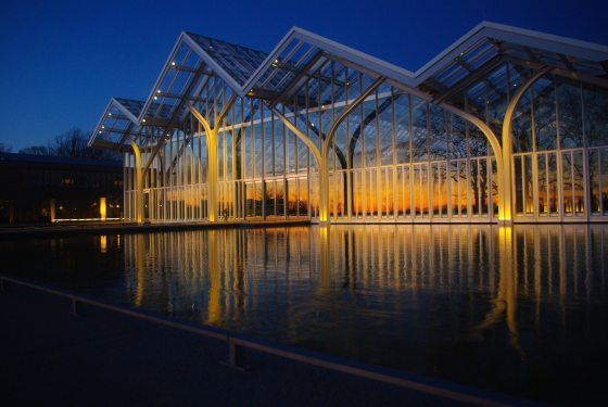 A glass conservatory, light up at night, with reflections of trees on it's facade.
